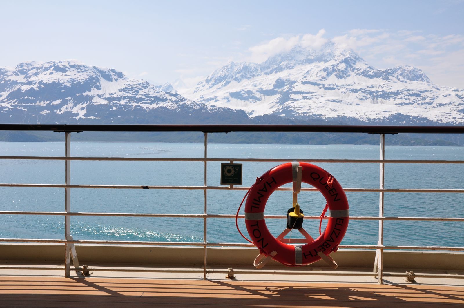 Snow-capped Alaska mountains seen over a ship’s rail and life ring, the view that makes the best time to book a cruise to Alaska matter